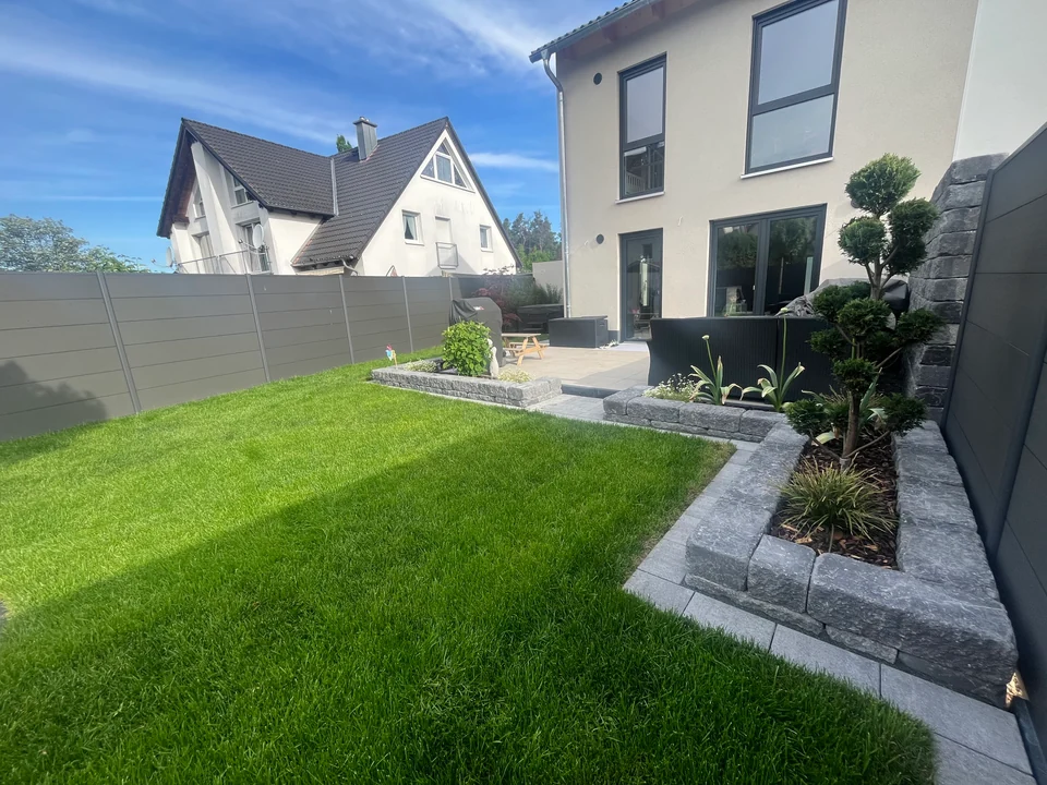 Hausgarten mit frischem Rasen, Hochbeeten mit Steinkanten und Terrasse hinter einem Neubau