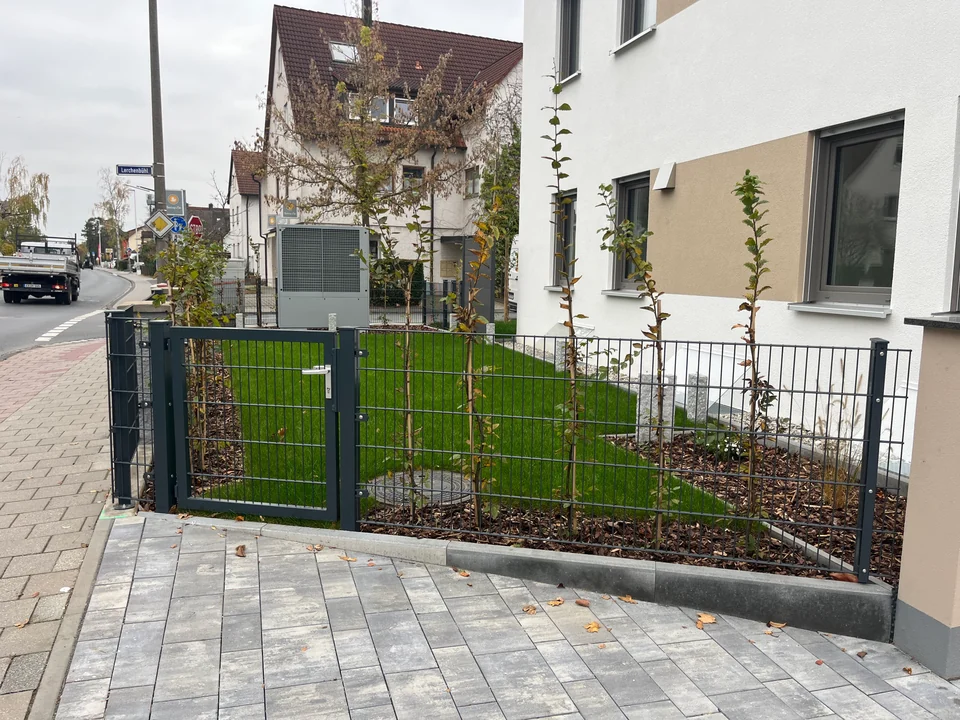 Neu angelegter Vorgarten mit Rasen, jungen Gehölzen und anthrazitfarbenem Stabmattenzaun mit Tor an einem Mehrfamilienhaus
