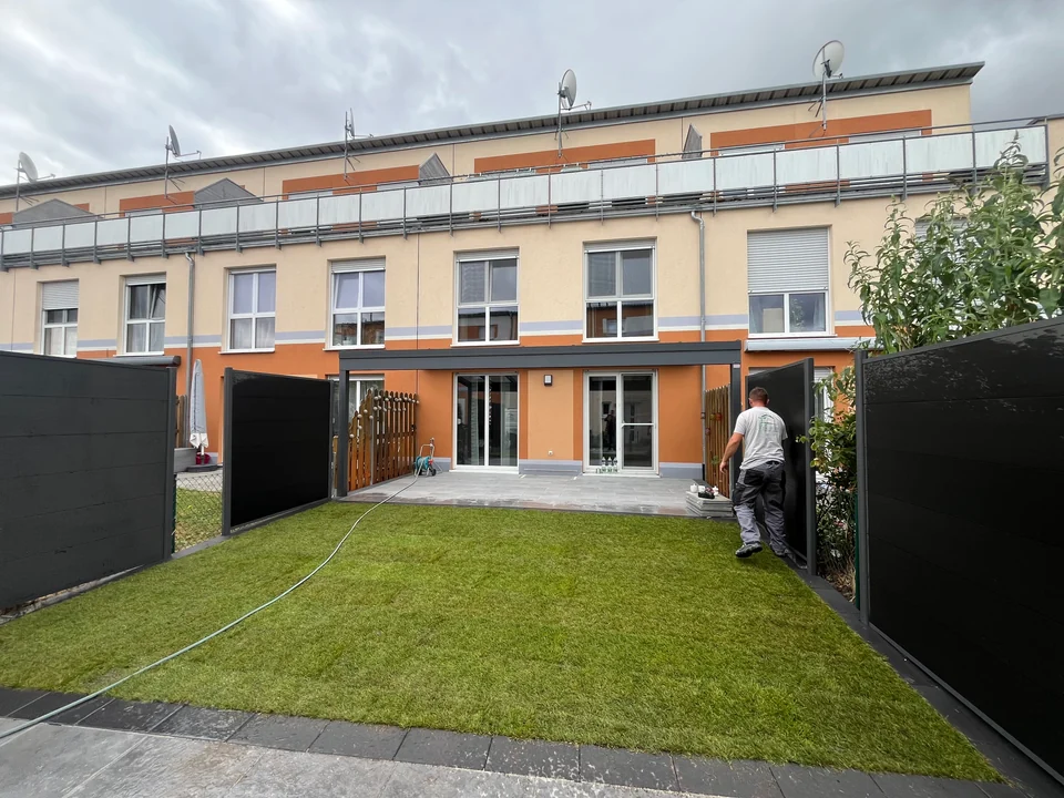 Reihenhausgarten mit frisch verlegtem Rasen, Terrasse und dunklen Sichtschutzelementen, ein Handwerker bei der Arbeit