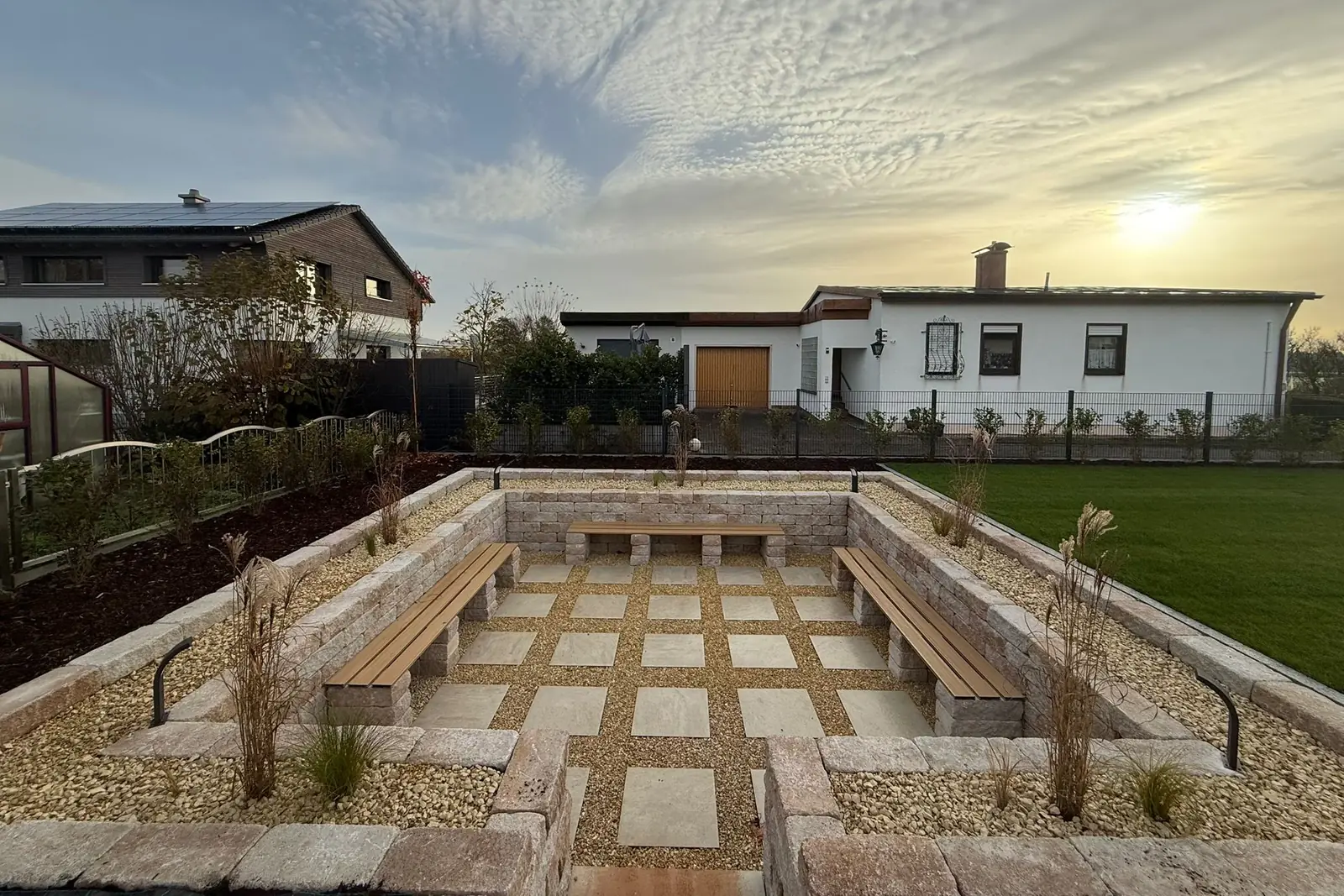 Abgesenkter Sitzplatz mit Natursteinmauern, drei Holzbänken auf Steinsockeln und Plattenraster in Kies