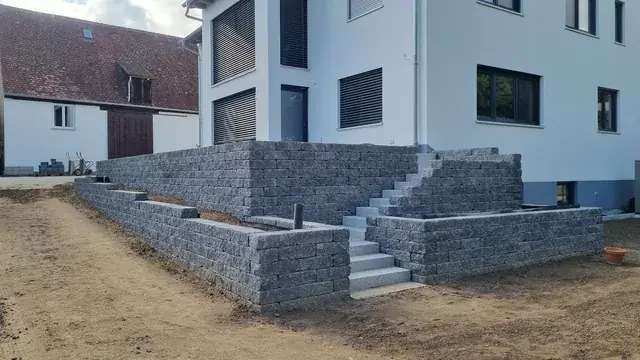 Terrassierte Mauern aus grauen Mauersteinen mit Treppe vor einem Neubau